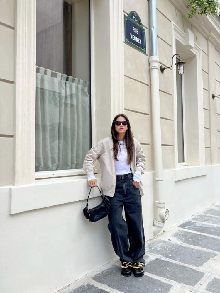 mulher usando calça jeans azul, blusa branca, jaqueta bege e sapato com corrente grossa. 