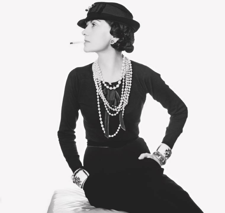 Coco Chanel sentada em seu ateliê em Paris, representando a filosofia de elegância e simplicidade